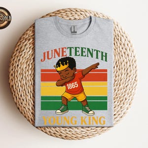 Puede incluir: Una camiseta gris con una ilustraci&oacute;n de dibujos animados de un ni&ntilde;o negro joven haciendo dab, con una corona y el n&uacute;mero 1865. La camiseta tiene las palabras "Juneteenth" y "Young King" en un dise&ntilde;o colorido.