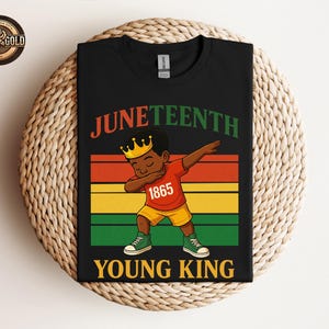 Puede incluir: Camiseta negra con un gr&aacute;fico colorido que celebra el Juneteenth. El dise&ntilde;o presenta una ilustraci&oacute;n de dibujos animados de un joven haciendo un dab, con una corona y el texto "Juneteenth Young King 1865". La camiseta se muestra sobre una superficie tejida.