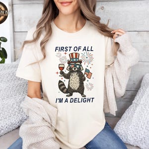 May include: Cream-colored t-shirt featuring a raccoon wearing sunglasses and a patriotic hat, holding a glass of wine and an American flag. The text "FIRST OF ALL I'M A DELIGHT" is printed above and below the raccoon illustration.