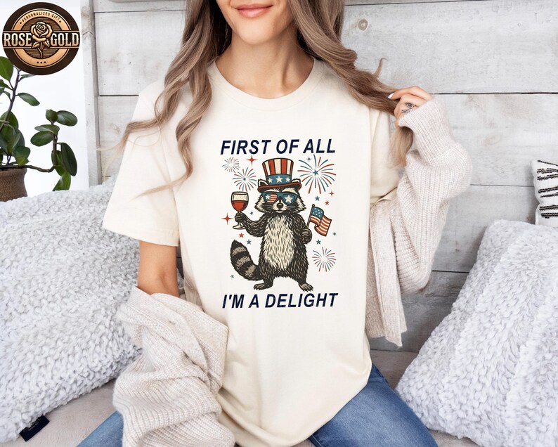 May include: Cream-colored t-shirt featuring a raccoon wearing sunglasses and a patriotic hat, holding a glass of wine and an American flag. The text "FIRST OF ALL I'M A DELIGHT" is printed above and below the raccoon illustration.