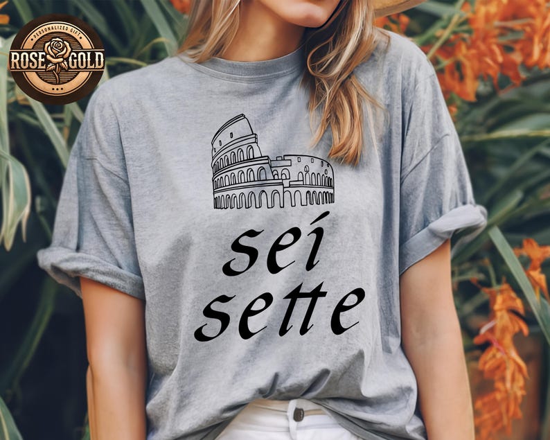 May include: A gray t-shirt featuring a black line drawing of the Colosseum and the Italian words "sei sette". The shirt has rolled-up sleeves and a relaxed fit. A rose gold logo is visible in the upper left corner.