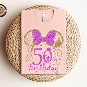 May include: A light pink t-shirt featuring a glittery purple bow and mouse ears design. The shirt has the text "50th Birthday" in a glittery purple font, with fireworks and a diamond graphic.