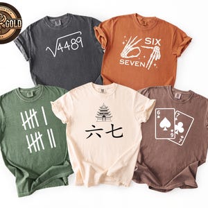 May include: Five different colored t-shirts with various graphic designs. The shirts are in shades of gray, orange, green, beige, and brown. Designs include a math equation, a skeleton hand, tally marks, a pagoda with Chinese characters, and playing cards.