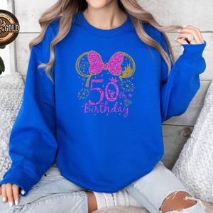 May include: Royal blue sweatshirt with a glittery pink bow and mouse ears design. The text "50th Birthday" is in pink glitter. Gold accents include a castle and fireworks. A rose gold logo is in the upper left corner.