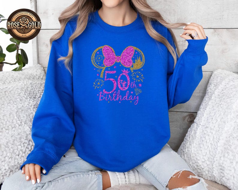 May include: Royal blue sweatshirt with a glittery pink bow and mouse ears design. The text "50th Birthday" is in pink glitter. Gold accents include a castle and fireworks. A rose gold logo is in the upper left corner.