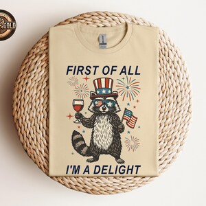 May include: Beige t-shirt featuring a raccoon wearing sunglasses and a patriotic hat, holding a glass of wine and an American flag. The text reads "FIRST OF ALL I'M A DELIGHT" in navy blue. The design includes fireworks and stars.