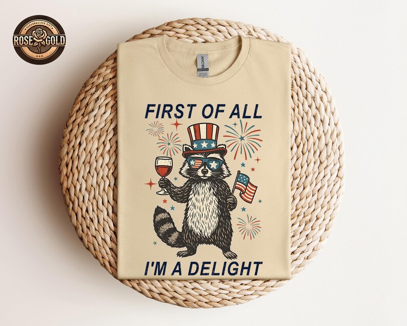May include: Beige t-shirt featuring a raccoon wearing sunglasses and a patriotic hat, holding a glass of wine and an American flag. The text reads "FIRST OF ALL I'M A DELIGHT" in navy blue. The design includes fireworks and stars.