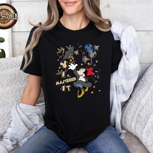 May include: Black t-shirt featuring a cartoon Mickey Mouse design with a red bow, yellow shoes, and the text "MASTERED IT." The design includes fireworks and stars. The shirt has a crew neck.