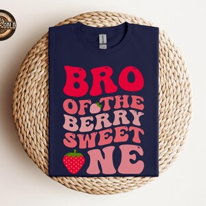 May include: Navy blue t-shirt with the text "BRO OF THE BERRY SWEET ONE" in red and pink lettering, with strawberry graphics. The shirt is displayed on a woven, circular surface.