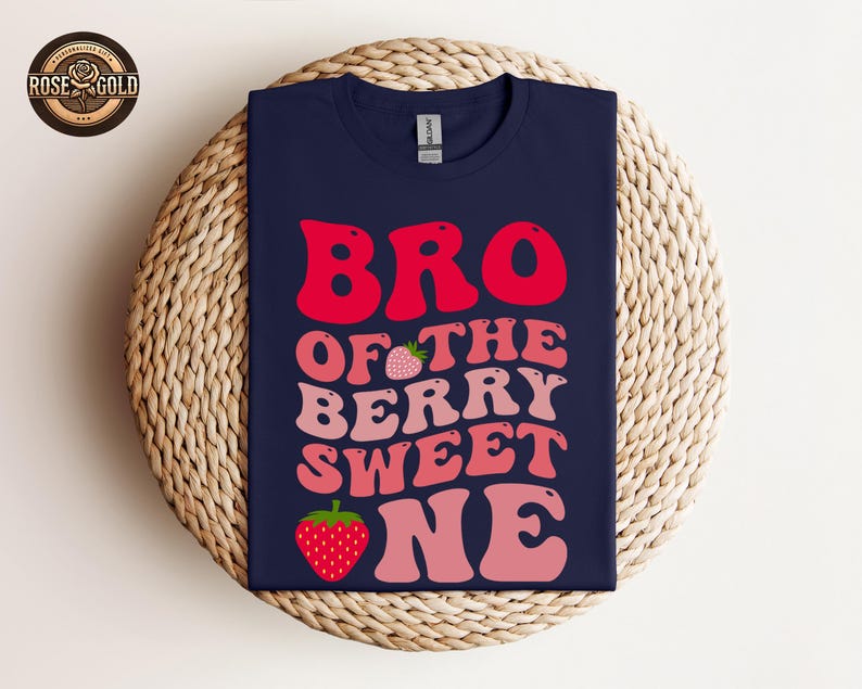 May include: Navy blue t-shirt with the text "BRO OF THE BERRY SWEET ONE" in red and pink lettering, with strawberry graphics. The shirt is displayed on a woven, circular surface.