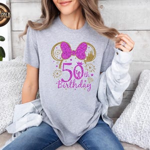 May include: A gray t-shirt with a glittery design celebrating a 50th birthday. The design includes a pink bow, gold mouse ears, a castle, fireworks, and the text "50th Birthday" in pink glitter.