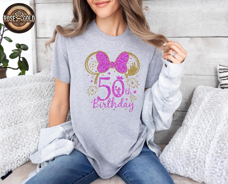 May include: A gray t-shirt with a glittery design celebrating a 50th birthday. The design includes a pink bow, gold mouse ears, a castle, fireworks, and the text "50th Birthday" in pink glitter.