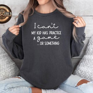 May include: Dark gray sweatshirt with the text "I can't, my kid has practice a game... or something" in white script. A rose gold logo is visible in the upper left corner. The sweatshirt is a casual style.