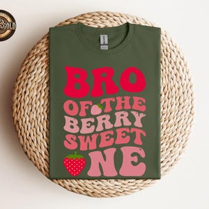 May include: An olive green t-shirt with the words "BRO OF THE BERRY SWEET ONE" in red and pink lettering, with strawberry graphics. The shirt is displayed on a woven, circular surface.