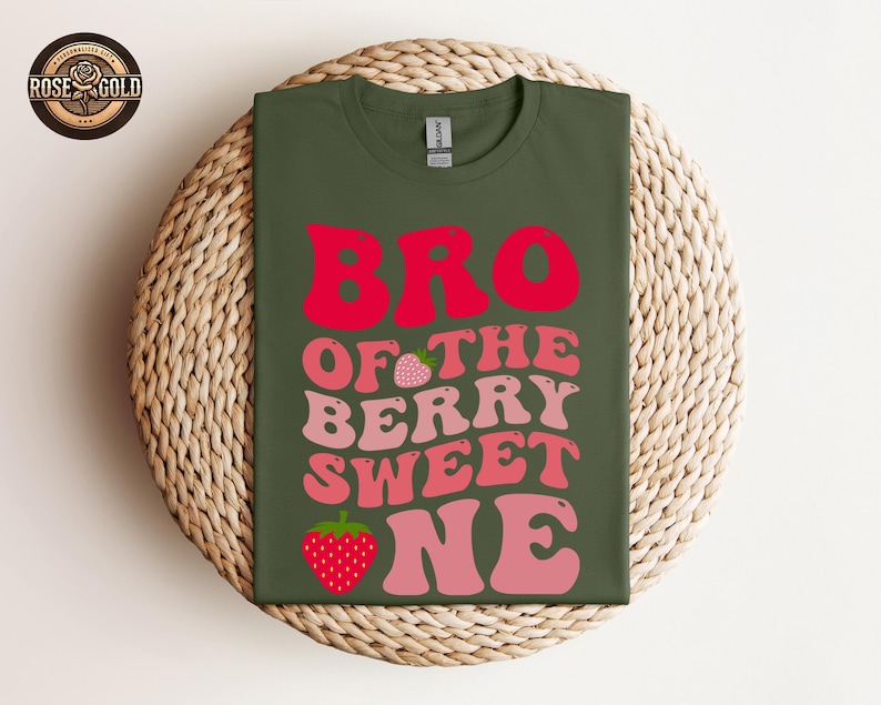 May include: An olive green t-shirt with the words "BRO OF THE BERRY SWEET ONE" in red and pink lettering, with strawberry graphics. The shirt is displayed on a woven, circular surface.