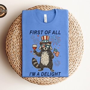 May include: A blue t-shirt featuring a raccoon wearing sunglasses, a patriotic hat, and holding a glass of wine and a small American flag. The text on the shirt reads "FIRST OF ALL I'M A DELIGHT".