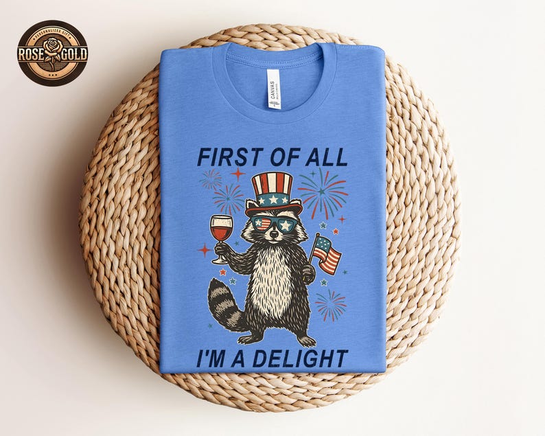 May include: A blue t-shirt featuring a raccoon wearing sunglasses, a patriotic hat, and holding a glass of wine and a small American flag. The text on the shirt reads "FIRST OF ALL I'M A DELIGHT".