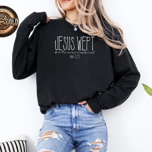 May include: Black long-sleeve sweatshirt with the text "JESUS WEPT" in white script. Below the main text is "but he never complained" and "John 11:35". A rose gold logo is in the upper left corner.