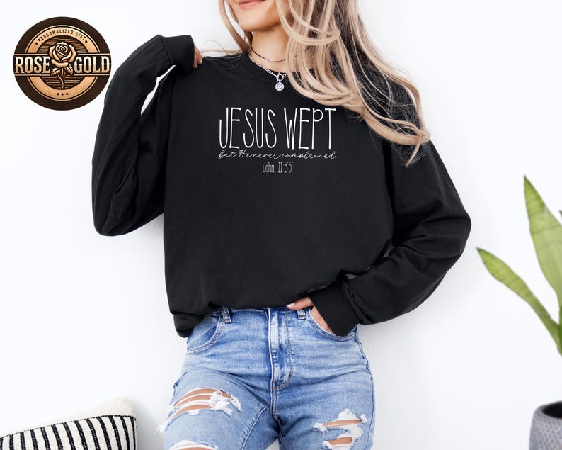 May include: Black long-sleeve sweatshirt with the text "JESUS WEPT" in white script. Below the main text is "but he never complained" and "John 11:35". A rose gold logo is in the upper left corner.