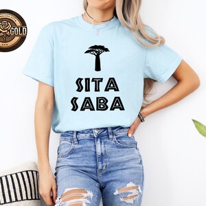 May include: Light blue t-shirt with a black tree graphic and the text "SITA SABA". The shirt is made of a soft material and is suitable for casual wear. The shirt is paired with blue jeans.