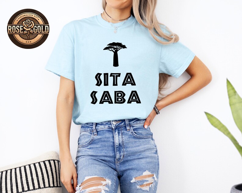 May include: Light blue t-shirt with a black tree graphic and the text "SITA SABA". The shirt is made of a soft material and is suitable for casual wear. The shirt is paired with blue jeans.