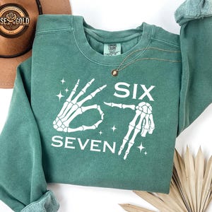 May include: A teal green sweatshirt with the words "SIX SEVEN" in white, accompanied by skeleton hand graphics. The sweatshirt has a crew neck and long sleeves. A gold necklace is draped on the front.