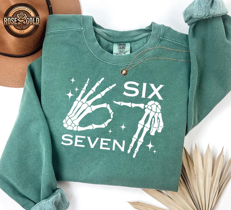 May include: A teal green sweatshirt with the words "SIX SEVEN" in white, accompanied by skeleton hand graphics. The sweatshirt has a crew neck and long sleeves. A gold necklace is draped on the front.