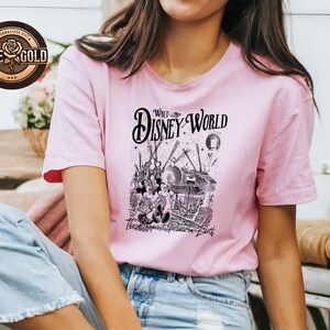 May include: A pale pink t-shirt with a black and white graphic of a Disney World scene. The text on the shirt reads "Walt Disney World" and "The Happiest Place on Earth". The t-shirt has short sleeves.
