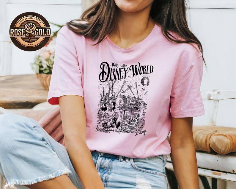 May include: A pale pink t-shirt with a black and white graphic of a Disney World scene. The text on the shirt reads "Walt Disney World" and "The Happiest Place on Earth". The t-shirt has short sleeves.
