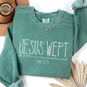 May include: A teal green sweatshirt with the text "JESUS WEPT" in white script. Below the text is "But He never complained" and "John 11:35". A gold necklace with a pendant is draped across the sweatshirt.