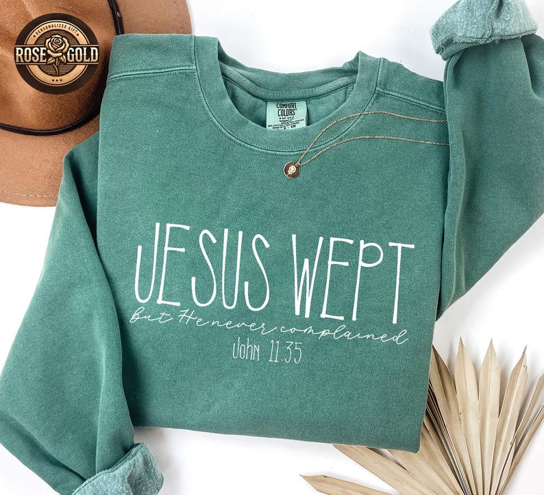 May include: A teal green sweatshirt with the text "JESUS WEPT" in white script. Below the text is "But He never complained" and "John 11:35". A gold necklace with a pendant is draped across the sweatshirt.