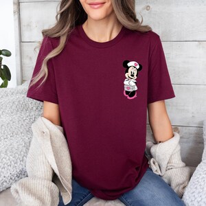 Puede incluir: Camiseta burdeos de cuello redondo con un gr&aacute;fico de Minnie Mouse. El gr&aacute;fico muestra a Minnie Mouse con uniforme de enfermera, sosteniendo una caja en forma de coraz&oacute;n. La camiseta es de color liso y tiene mangas cortas.