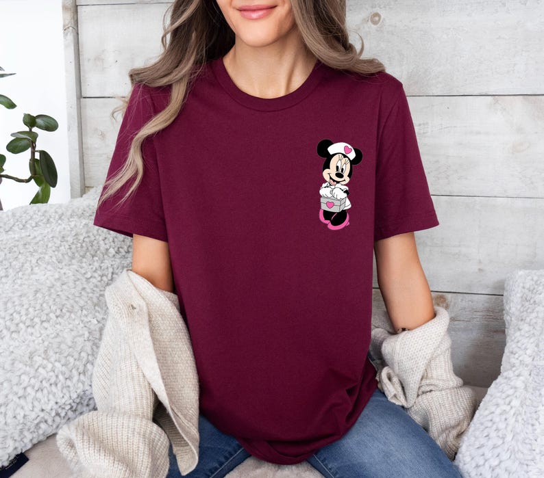 Puede incluir: Camiseta burdeos de cuello redondo con un gr&aacute;fico de Minnie Mouse. El gr&aacute;fico muestra a Minnie Mouse con uniforme de enfermera, sosteniendo una caja en forma de coraz&oacute;n. La camiseta es de color liso y tiene mangas cortas.