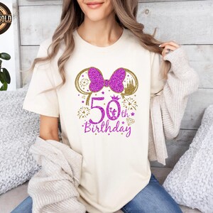 May include: Cream-colored t-shirt with a glittery purple bow and mouse ears design. The shirt features the text "50th Birthday" in purple, with gold accents. A castle and fireworks complete the design.