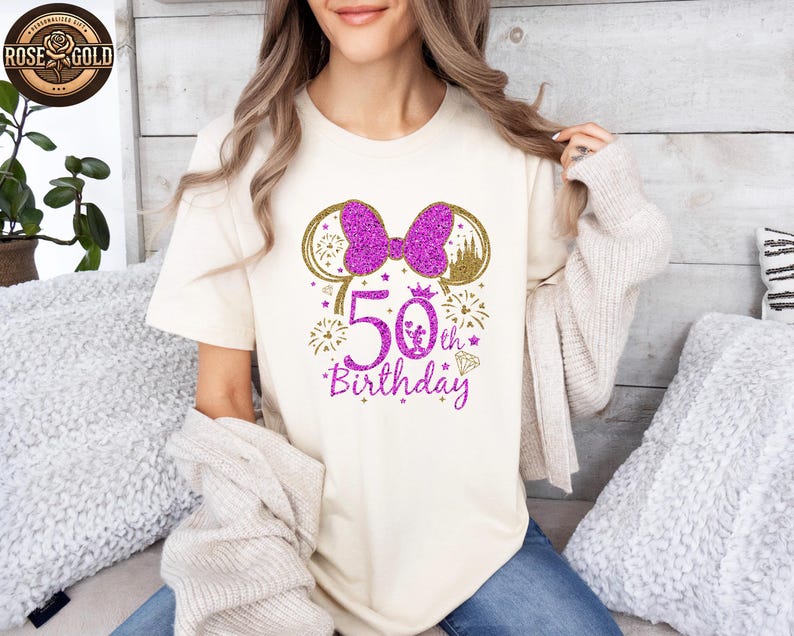May include: Cream-colored t-shirt with a glittery purple bow and mouse ears design. The shirt features the text "50th Birthday" in purple, with gold accents. A castle and fireworks complete the design.