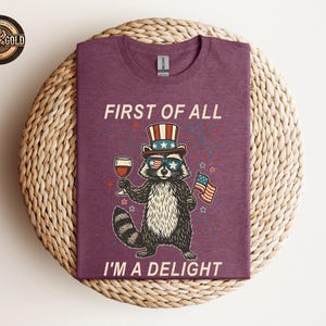 May include: A maroon t-shirt featuring a raccoon wearing sunglasses, a patriotic hat, and holding a glass of wine and an American flag. The text "FIRST OF ALL I'M A DELIGHT" is printed on the shirt.