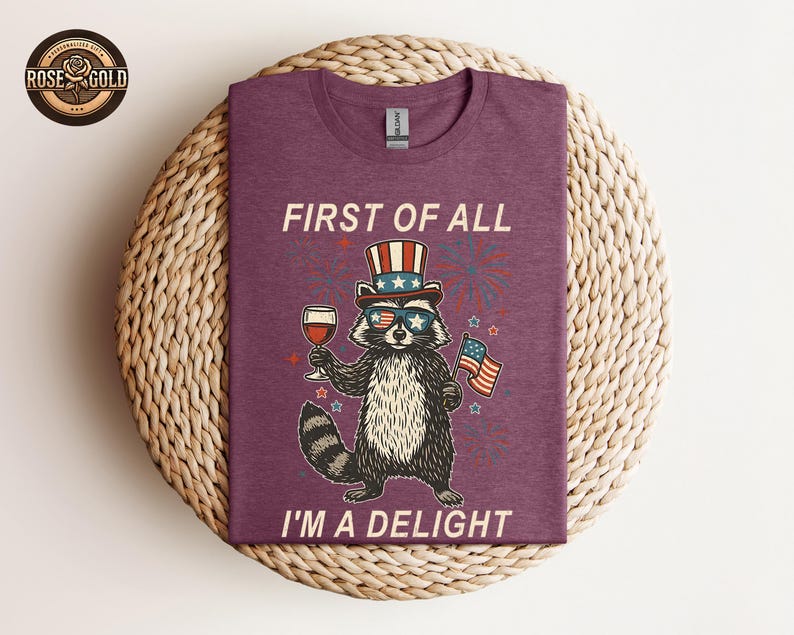 May include: A maroon t-shirt featuring a raccoon wearing sunglasses, a patriotic hat, and holding a glass of wine and an American flag. The text "FIRST OF ALL I'M A DELIGHT" is printed on the shirt.