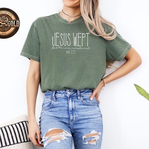 May include: An olive green t-shirt with the text "JESUS WEPT" in a white, handwritten font. Below the text is "but He never complained" and "John 11:35". The shirt is paired with blue jeans.