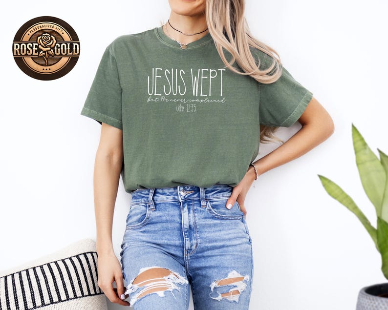 May include: An olive green t-shirt with the text "JESUS WEPT" in a white, handwritten font. Below the text is "but He never complained" and "John 11:35". The shirt is paired with blue jeans.