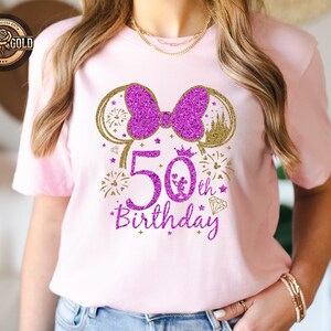 May include: A light pink t-shirt with a glittery purple design celebrating a 50th birthday. The design includes a bow, mouse ears, fireworks, a crown, and the words "50th Birthday". The shirt is a short-sleeved crew neck.