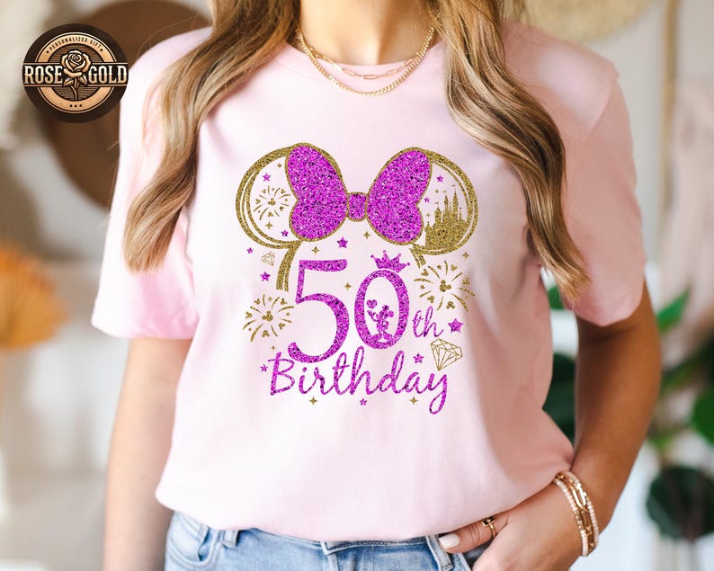 May include: A light pink t-shirt with a glittery purple design celebrating a 50th birthday. The design includes a bow, mouse ears, fireworks, a crown, and the words "50th Birthday". The shirt is a short-sleeved crew neck.