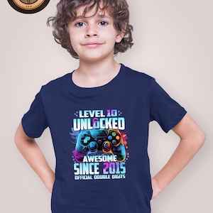 May include: Navy blue t-shirt with a colorful graphic of a video game controller and the text "LEVEL 10 UNLOCKED AWESOME SINCE 2015 OFFICIAL DOUBLE DIGITS". The shirt also has a "ROSE GOLD" logo.