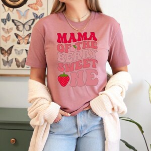 May include: A dusty rose t-shirt with the text "MAMA OF THE BERRY SWEET ONE" in shades of pink. The design includes strawberry graphics. The shirt is short-sleeved and worn with light blue jeans and a cream-colored cardigan.