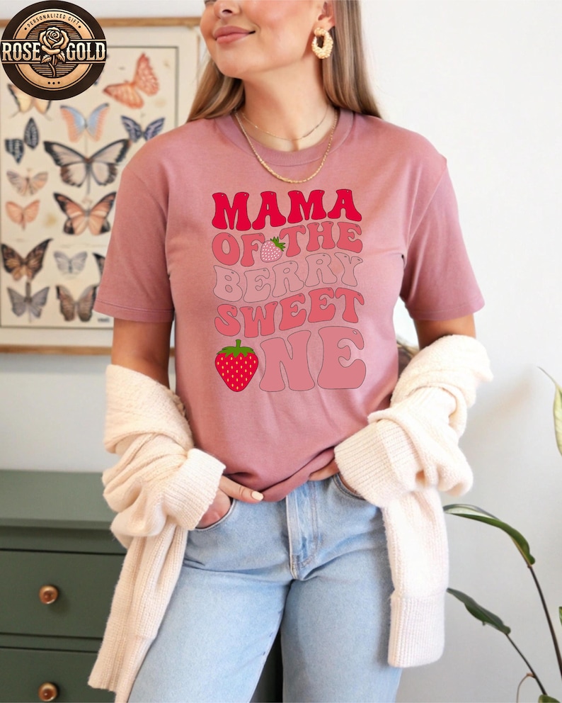 May include: A dusty rose t-shirt with the text "MAMA OF THE BERRY SWEET ONE" in shades of pink. The design includes strawberry graphics. The shirt is short-sleeved and worn with light blue jeans and a cream-colored cardigan.