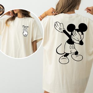 May include: Cream-coloured t-shirt with a black and white Mickey Mouse graphic on the back, covering his eyes. The front of the shirt has the word "Taylor" and a hand gesture.