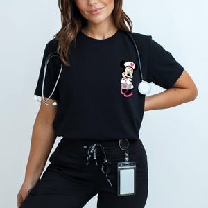 Puede incluir: Camiseta negra con un dise&ntilde;o de Minnie Mouse enfermera. El personaje de dibujos animados lleva un gorro y un uniforme de enfermera. Un estetoscopio est&aacute; alrededor del cuello. La camiseta se combina con pantalones negros y un cord&oacute;n.