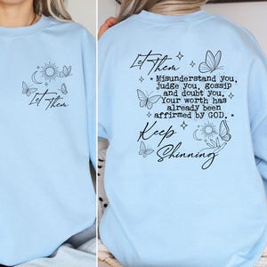 May include: Light blue sweatshirt with a black graphic print of butterflies, stars, and the text "Let Them" and "Keep Shining" with a quote about worth being affirmed by God.