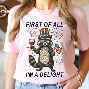 May include: Light pink t-shirt featuring a raccoon wearing sunglasses, a patriotic hat, and holding a glass of wine and an American flag. The text on the shirt reads, "FIRST OF ALL I'M A DELIGHT."