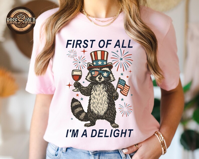 May include: Light pink t-shirt featuring a raccoon wearing sunglasses, a patriotic hat, and holding a glass of wine and an American flag. The text on the shirt reads, "FIRST OF ALL I'M A DELIGHT."