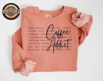 Puede incluir: Una sudadera de color coral con las palabras "Coffee Addict" en una fuente negra. La camisa también presenta una lista de frases relacionadas con el café. Una scrunchie a juego descansa sobre la manga. Un logotipo de oro rosa está en la esquina superior izquierda.